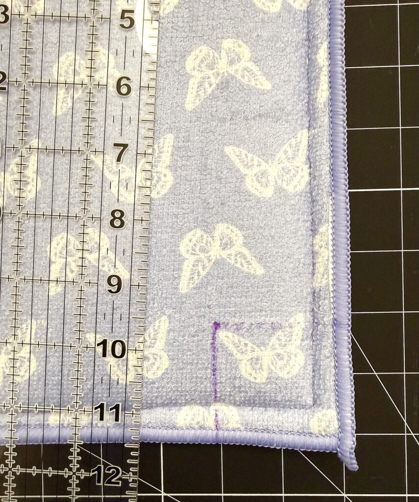 A clear ruler is placed on a fabric with white butterfly prints and purple edges, next to a marked cutting mat.