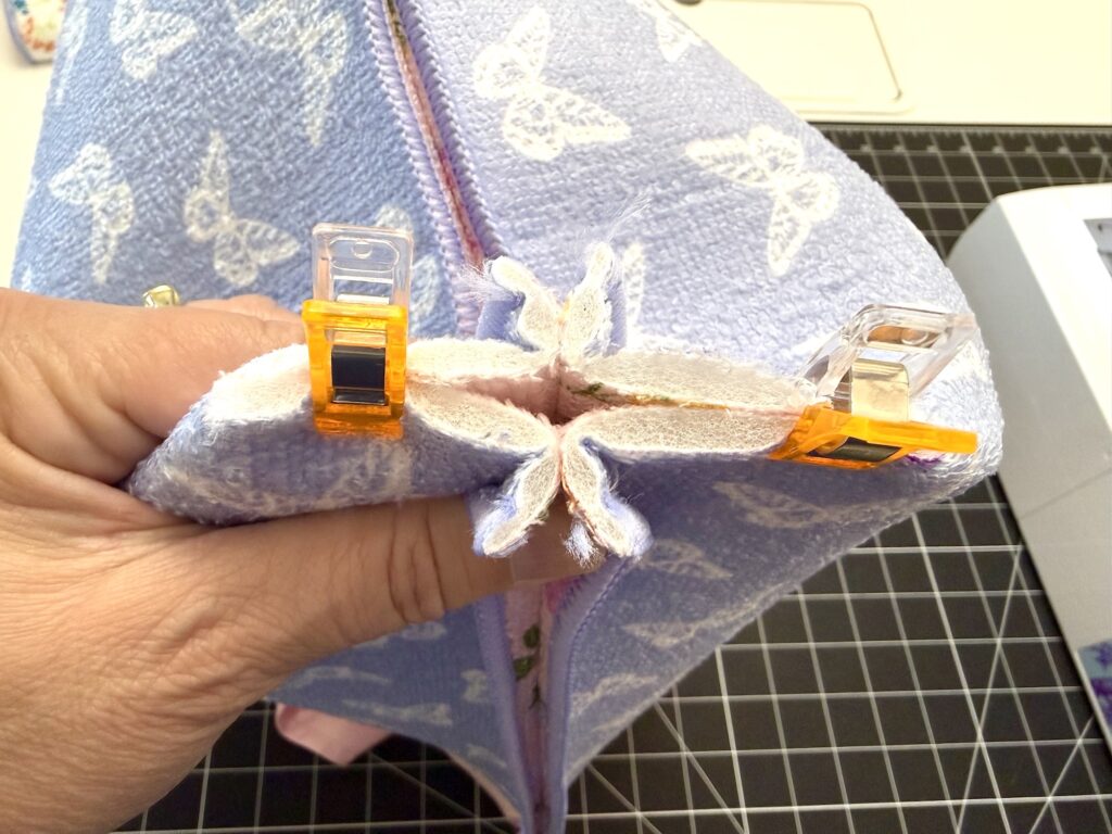 A hand holds fabric pieces clipped together with orange and clear clips, preparing to sew along the edge; a sewing machine and grid cutting mat are visible.