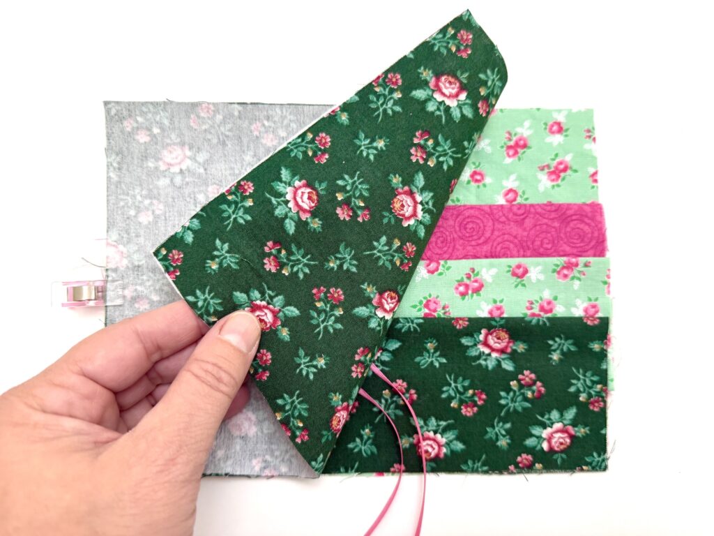A hand lifts a green floral fabric piece to reveal layered fabric strips beneath, including floral and solid designs, on a white background—ideal for crafting a charming tea wallet.