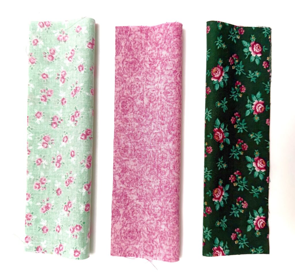Three rectangular fabric swatches—a light green with pink flowers, solid pink, and dark green with red and pink floral print—are arranged vertically on a white background, perfect for crafting a charming tea wallet.