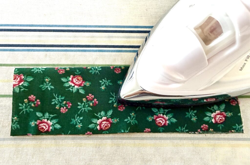 An iron presses a piece of green floral fabric on a striped ironing board, perfect for crafting a charming tea wallet.