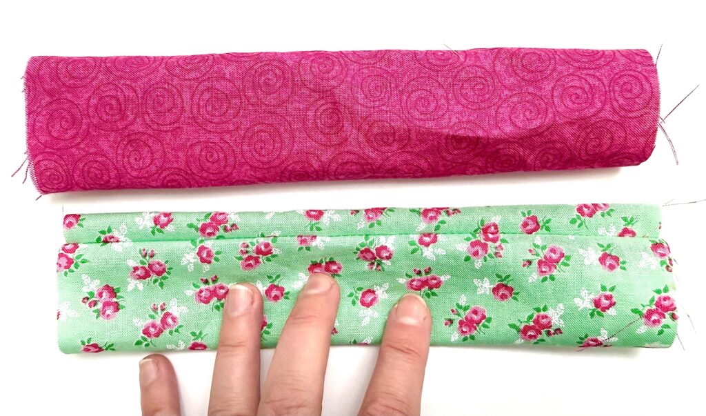 Two pieces of fabric on a white surface: one pink with swirls, rolled up; one green with pink flower print, partially folded and held by three fingers—perfect for crafting a charming tea wallet.