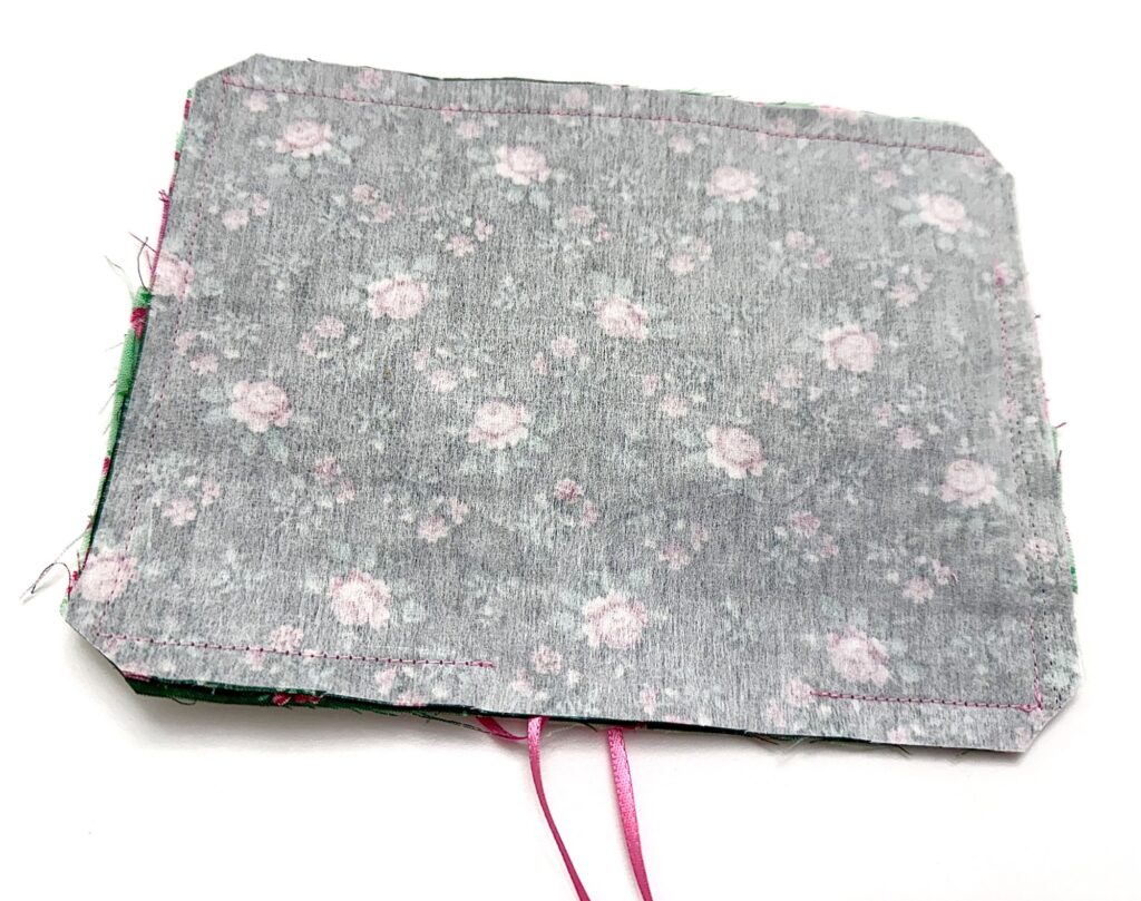 A rectangular tea wallet with a floral pattern is sewn along the edges with pink thread; two pink ribbons extend from the bottom.