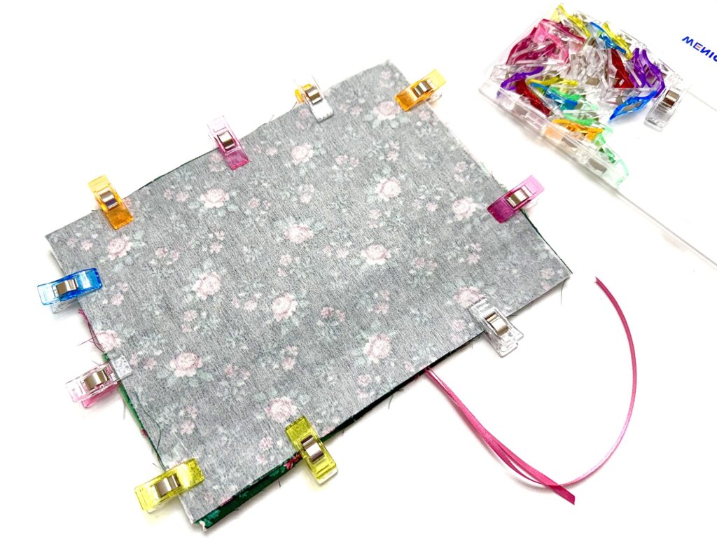 A piece of floral fabric for a tea wallet is held by colorful sewing clips on the edges, with a pink ribbon underneath and more clips in a clear box nearby.