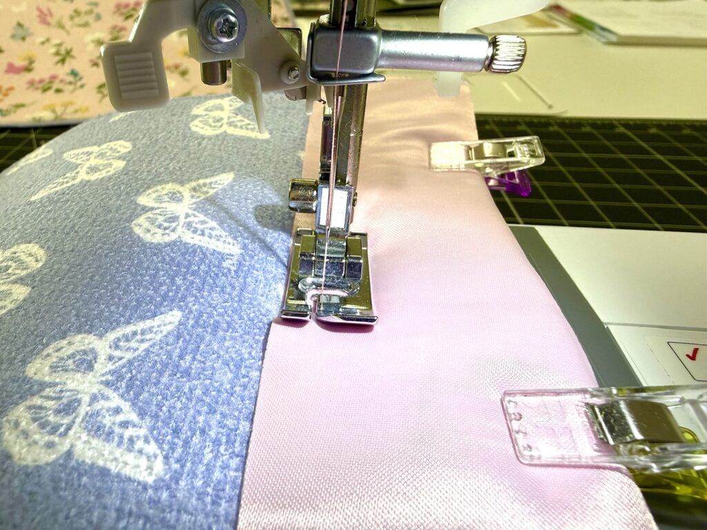 Close-up of a sewing machine stitching together blue patterned and pink fabrics, held in place by clips.