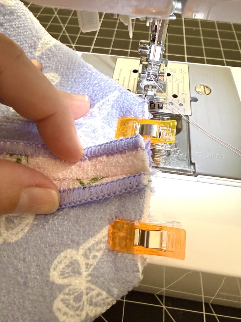 A person uses orange sewing clips to hold fabric pieces together while positioning them under a sewing machine needle for stitching.