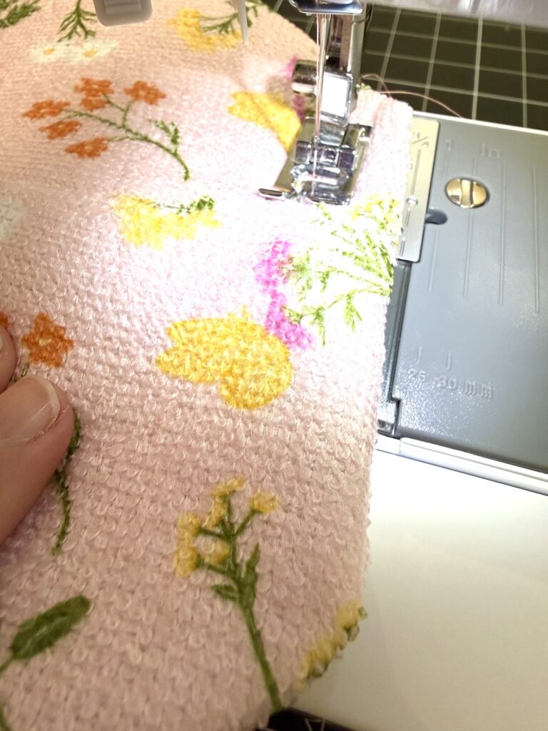 A close-up of a sewing machine stitching pink, textured fabric with a floral pattern; a hand is guiding the fabric under the needle.