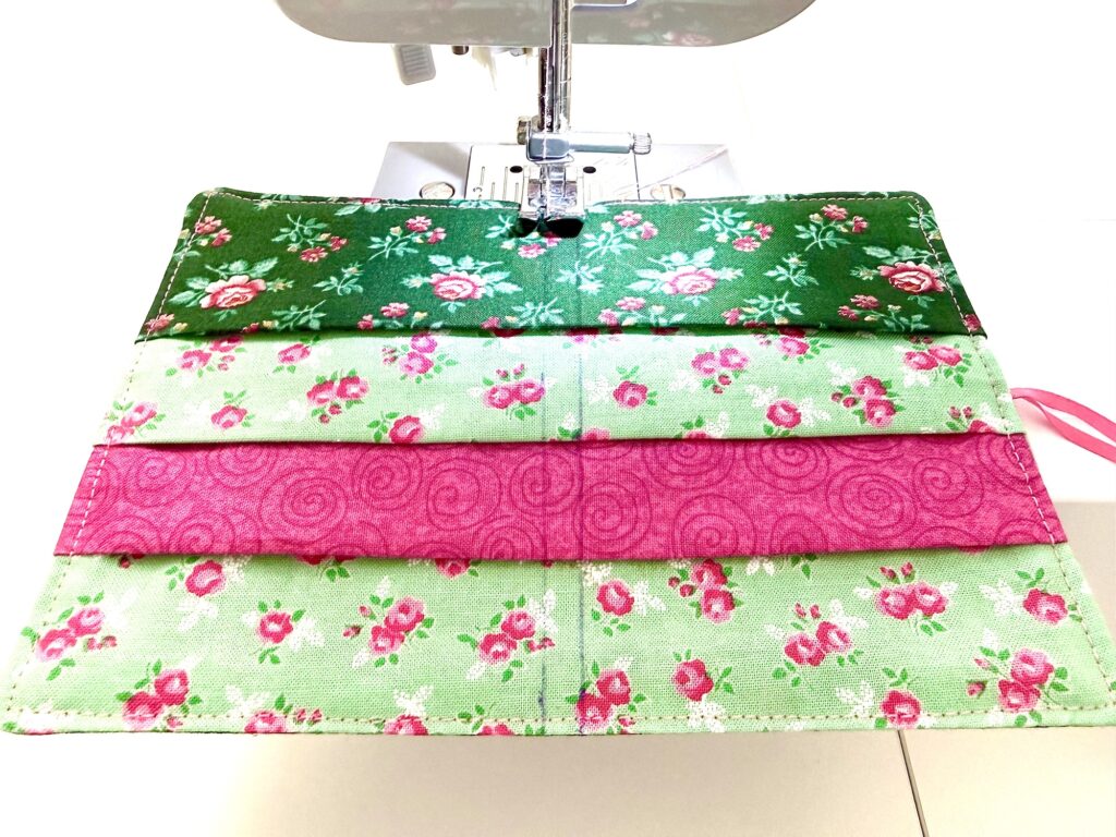 A sewing machine stitching a fabric piece with green and pink floral patterns and horizontal pockets, perfect for creating a charming tea wallet.