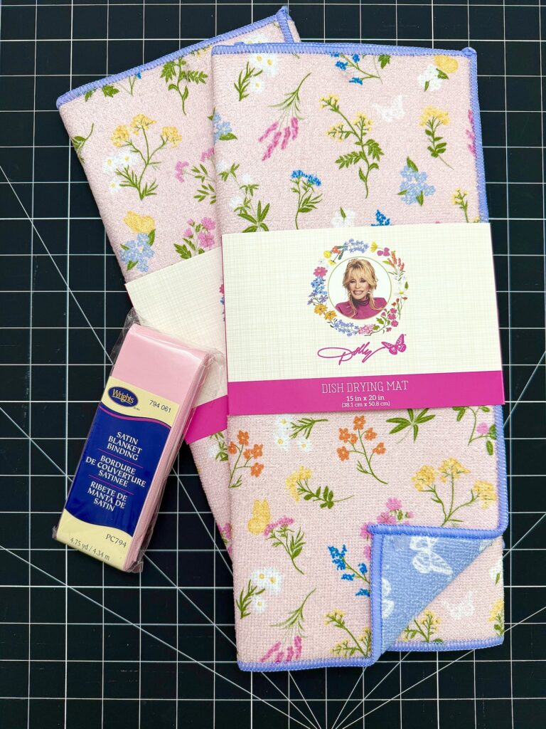 A pack of pink satin ribbon and a folded floral dish drying mat with a portrait of Dolly Parton on the packaging, placed on a black grid surface.
