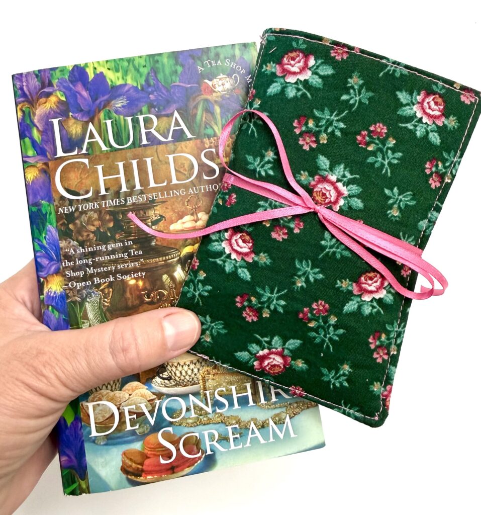 A hand holds a book titled "Devonshire Scream" by Laura Childs and a green floral tea wallet case tied with a pink ribbon.