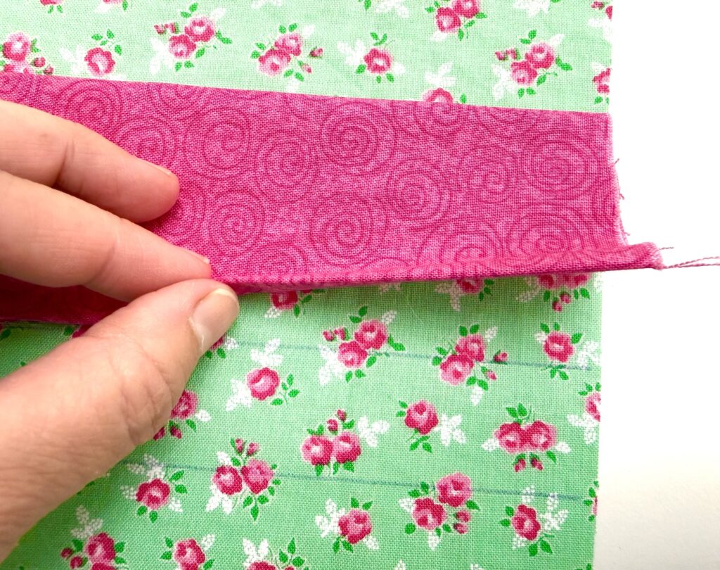 A hand holds the edge of a pink strip of fabric sewn onto green floral fabric, showing the seam and pattern details—perfect for crafting a unique tea wallet.