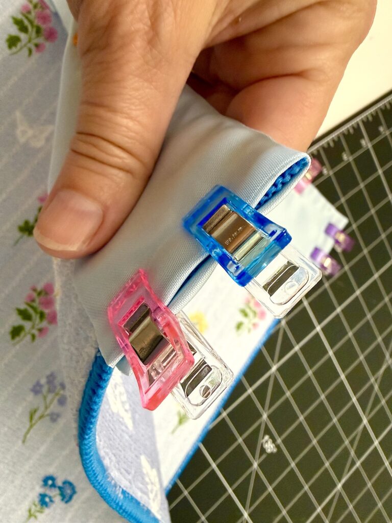 A hand holds fabric layers secured with pink and blue sewing clips for a dish mat tote bag, positioned on a cutting mat with floral fabric visible underneath.