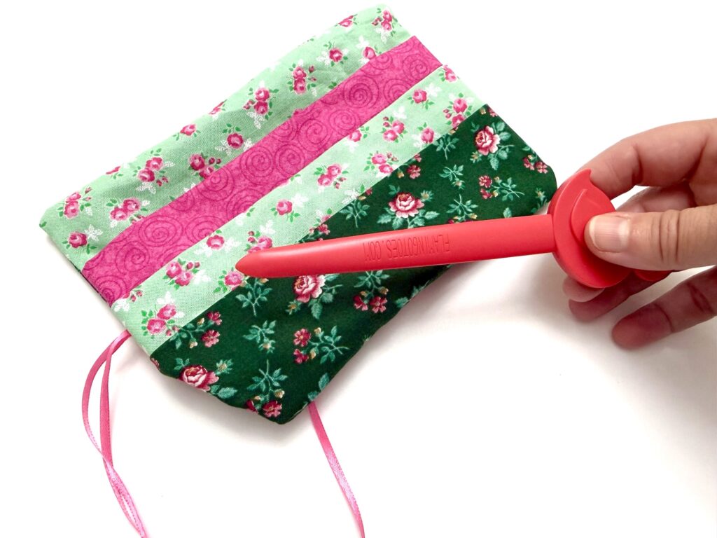 A hand holds a red plastic tool next to a small, colorful floral fabric tea wallet with pink and green patterns and ribbon ties.