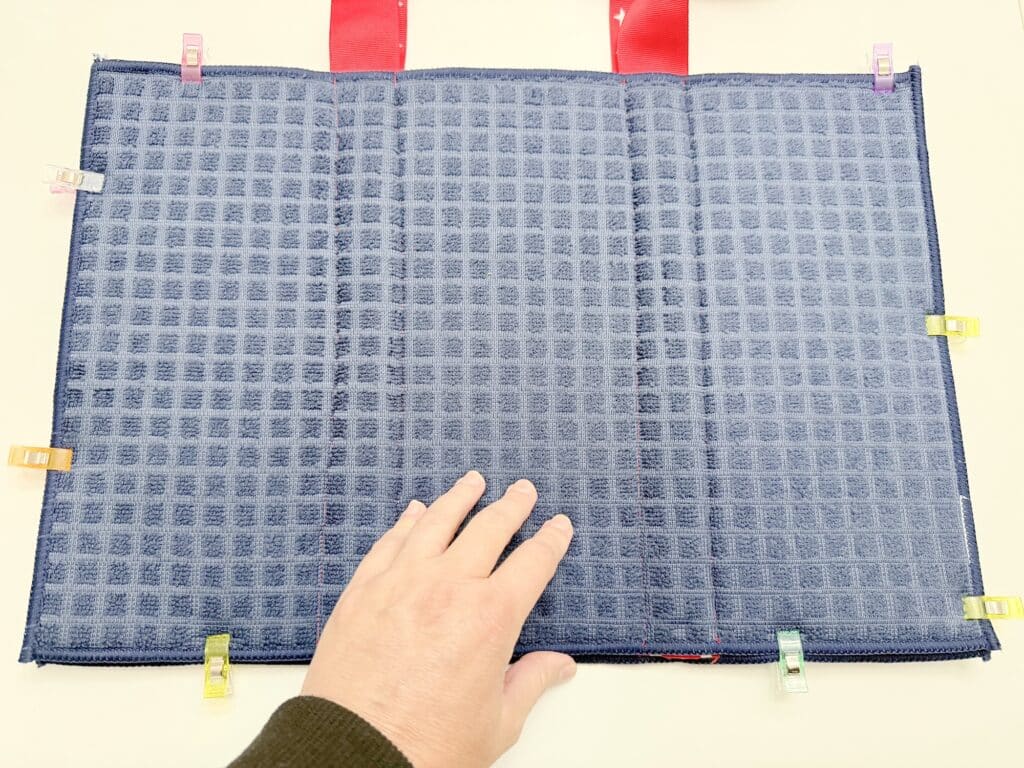 A hand rests on a dark blue textured fabric secured with colorful clips along the edges; two red ribbons are visible at the top.