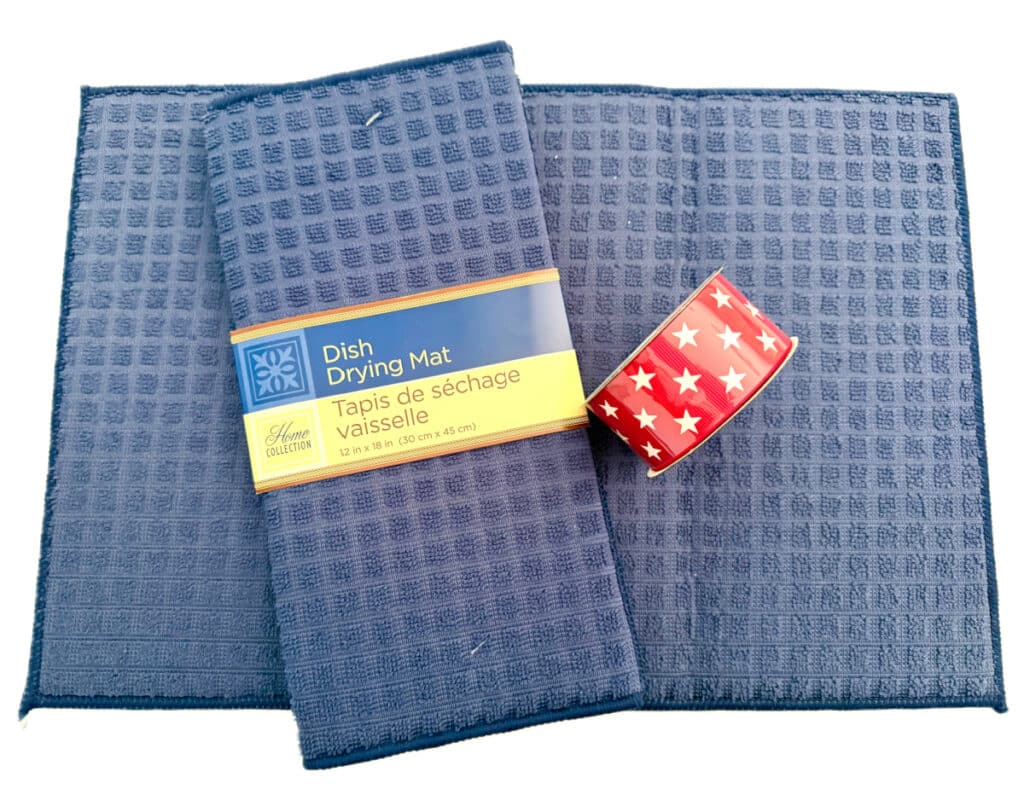Two blue dish drying mats, one folded with a label, with a roll of red tape featuring white stars on top—perfect for storing together in a dish mat bag.