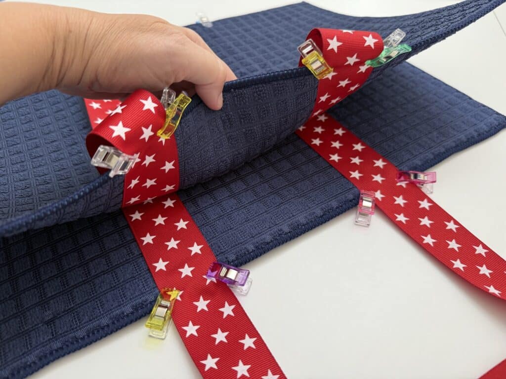 A hand holds blue textured fabric, while red ribbon with white stars and colorful clips are attached, likely in preparation for sewing a dish mat bag or crafting another creative project.