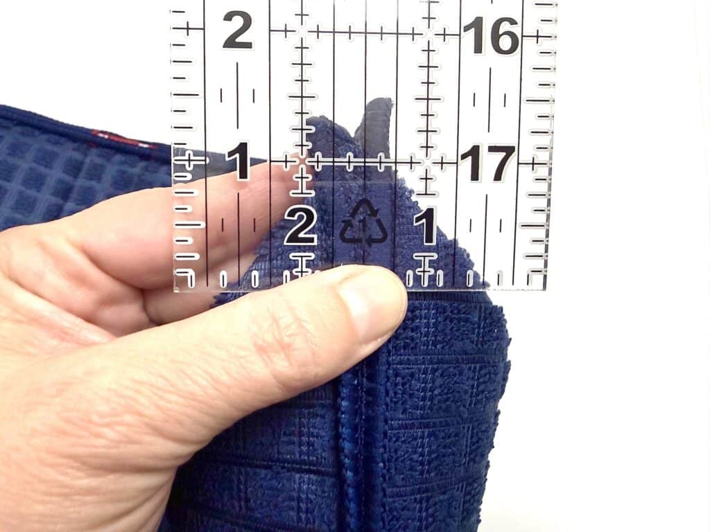 A hand holds a ruler vertically against the seam of a folded blue textured fabric, measuring the thickness at approximately 2 centimeters.