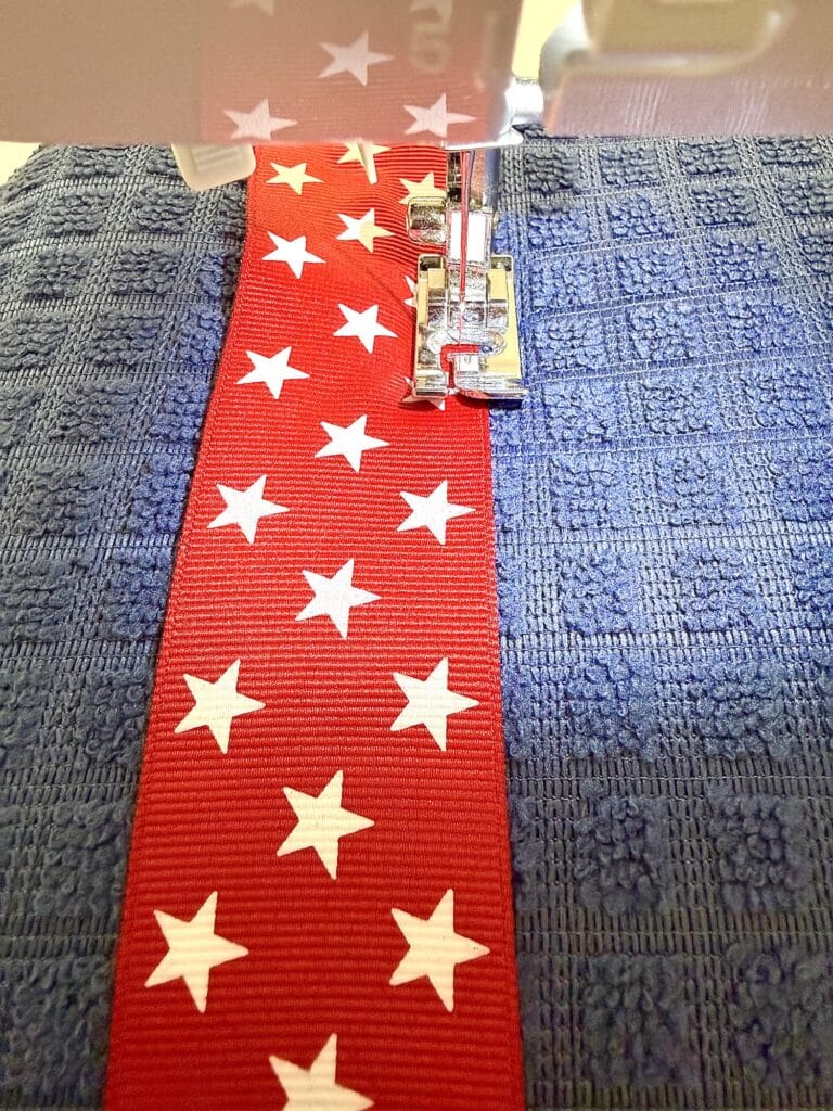 A sewing machine stitches a red ribbon with white stars onto a textured blue fabric.