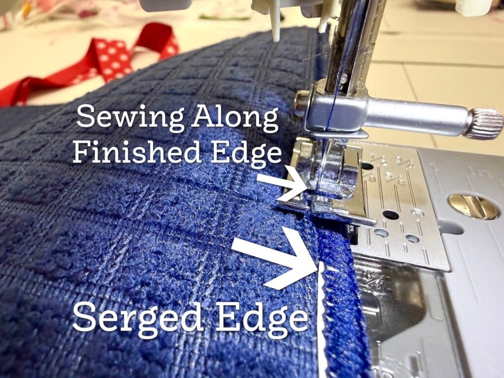 Close-up of a sewing machine stitching along a blue fabric edge, with text labeling the finished edge being sewn and the previously serged edge.