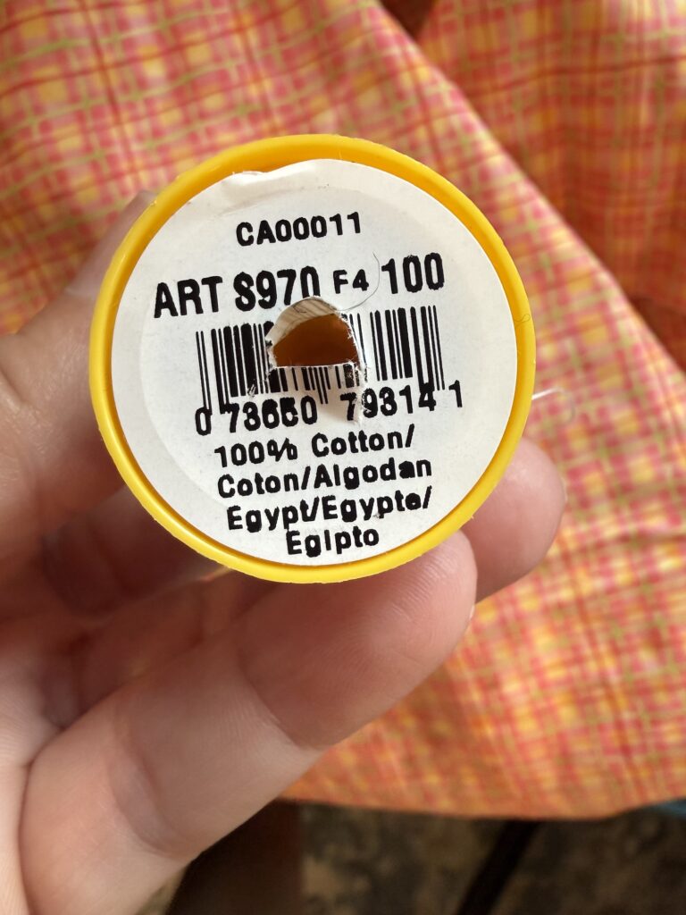 A hand holds a yellow spool of thread labeled "100% Cotton, Egypt" with a barcode and product details printed on the end.