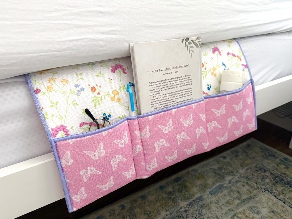 Pink bedside caddy with white butterfly pattern holds a book, glasses, a pen, and other small items; tucked under a bed with floral sheets.