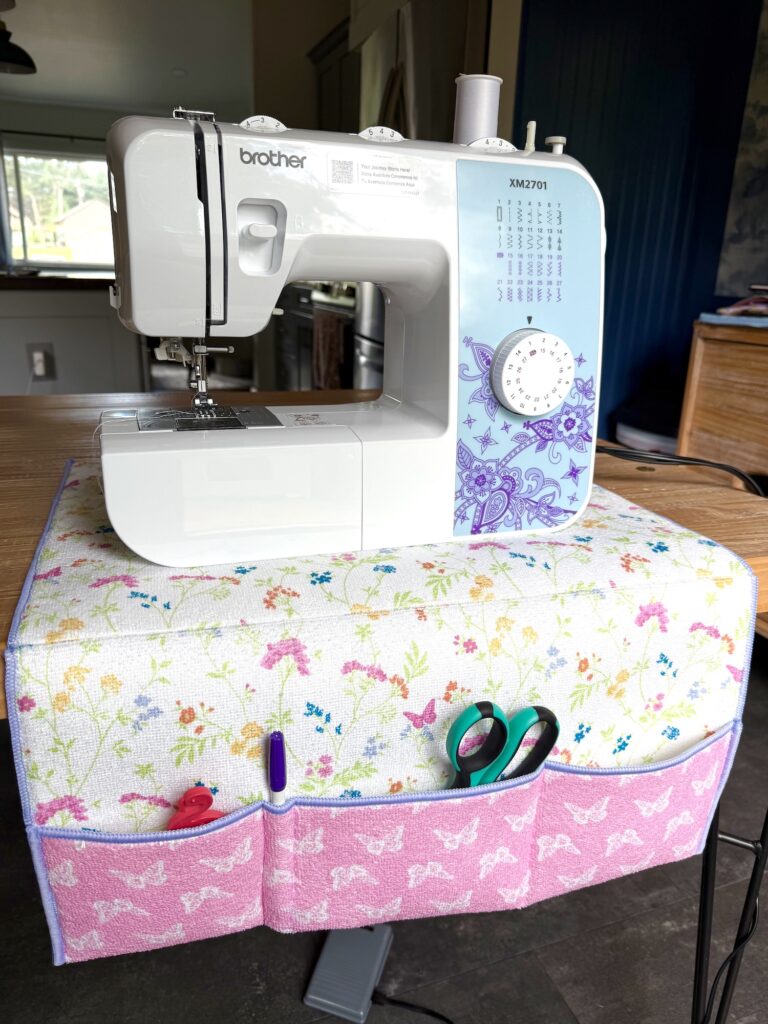 A Brother sewing machine sits on a table, covered with a floral and pink mat holding scissors, pens, and other sewing tools in front-facing pockets.