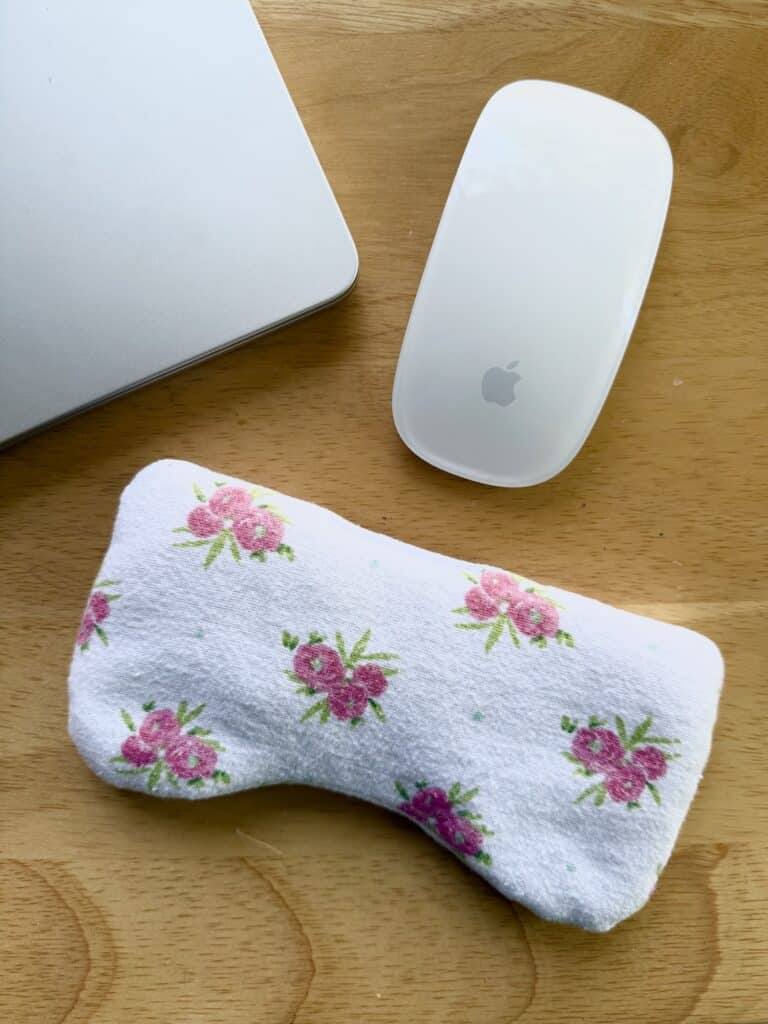 A white computer Mouse, part of a silver laptop, and a white floral fabric mouse wrist rest are on a wooden desk.