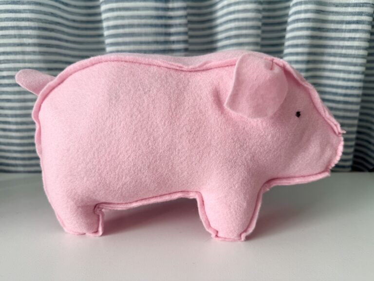A handmade pink felt pig plush stands on a white surface, with blue and white striped fabric in the background.