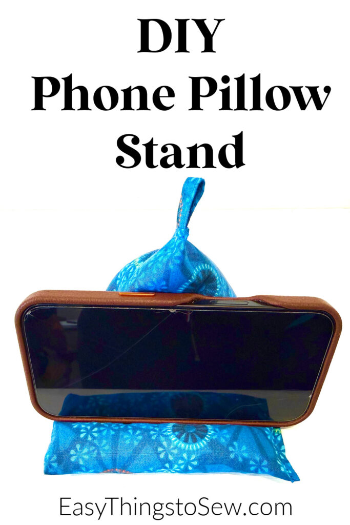 A smartphone rests on a blue, fabric phone pillow stand with a small handle, displaying the text "DIY Phone Pillow Stand" and "EasyThingsToSew.com" above and below.