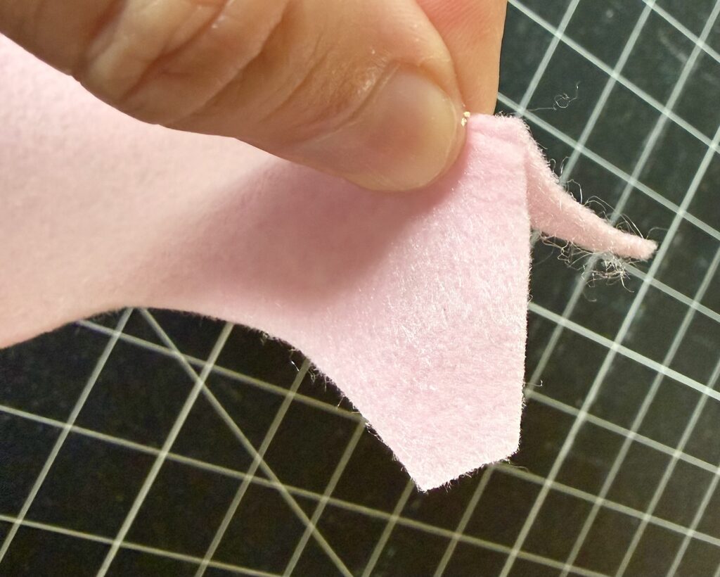 A hand holding the edge of a piece of light pink felt, with a grid-patterned cutting mat in the background.