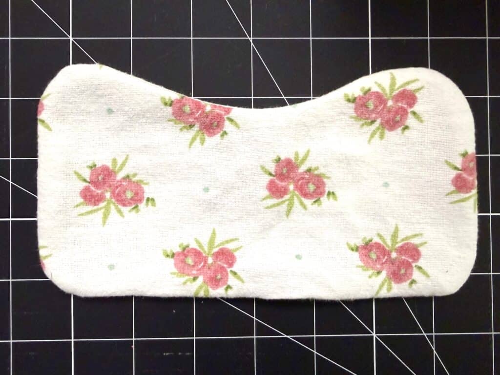 A rectangular fabric pad with curved corners and a floral pattern featuring pink flowers on a white background, placed on a black grid cutting mat.