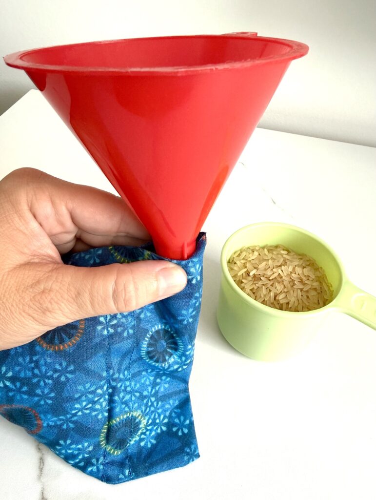 A hand holds a red funnel over a blue fabric pouch, with a green measuring cup of uncooked rice beside them on a white surface.