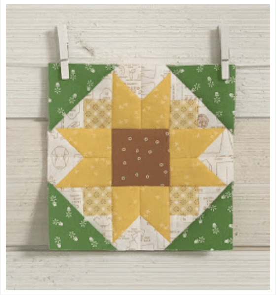 A quilt block featuring a yellow and brown sunflower design with green corners, displayed against a white wooden background.