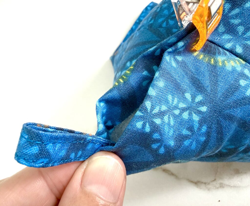 A hand holds a blue fabric loop attached to a patterned, sewn item, secured with an orange sewing clip on a white surface.