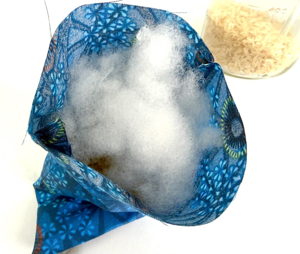 Open blue fabric pouch filled with white stuffing material, with a jar of uncooked rice visible in the background on a white surface.