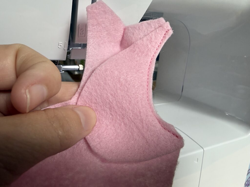 A hand holds pink fabric with a curved seam next to a sewing machine needle plate, showing sewing in progress.