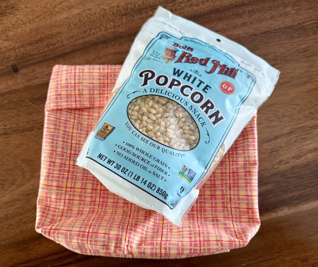 A bag of Bob's Red Mill white popcorn sits on top of a pink and yellow checkered cloth on a wooden surface.