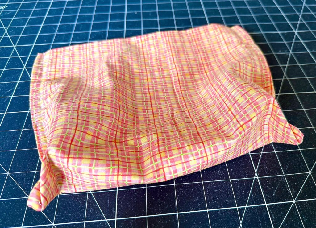 A pink and yellow plaid fabric face mask is placed on a black gridded cutting mat.