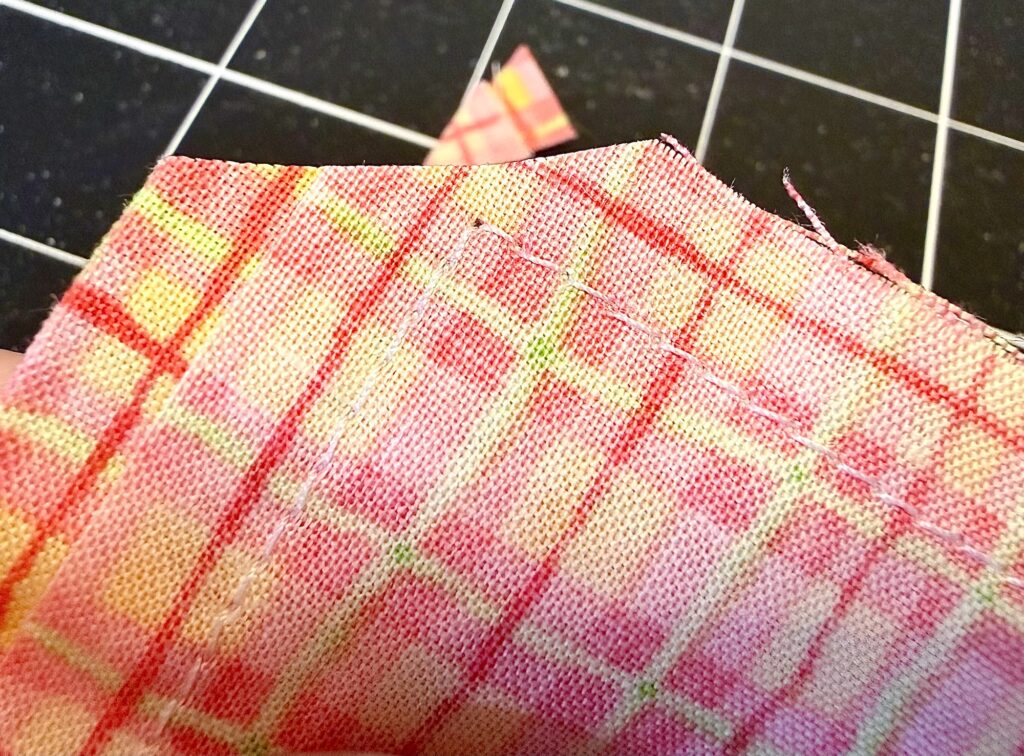 A close-up of a piece of pink and yellow plaid fabric showing a line of hand stitching along the edge, with a grid cutting mat in the background.