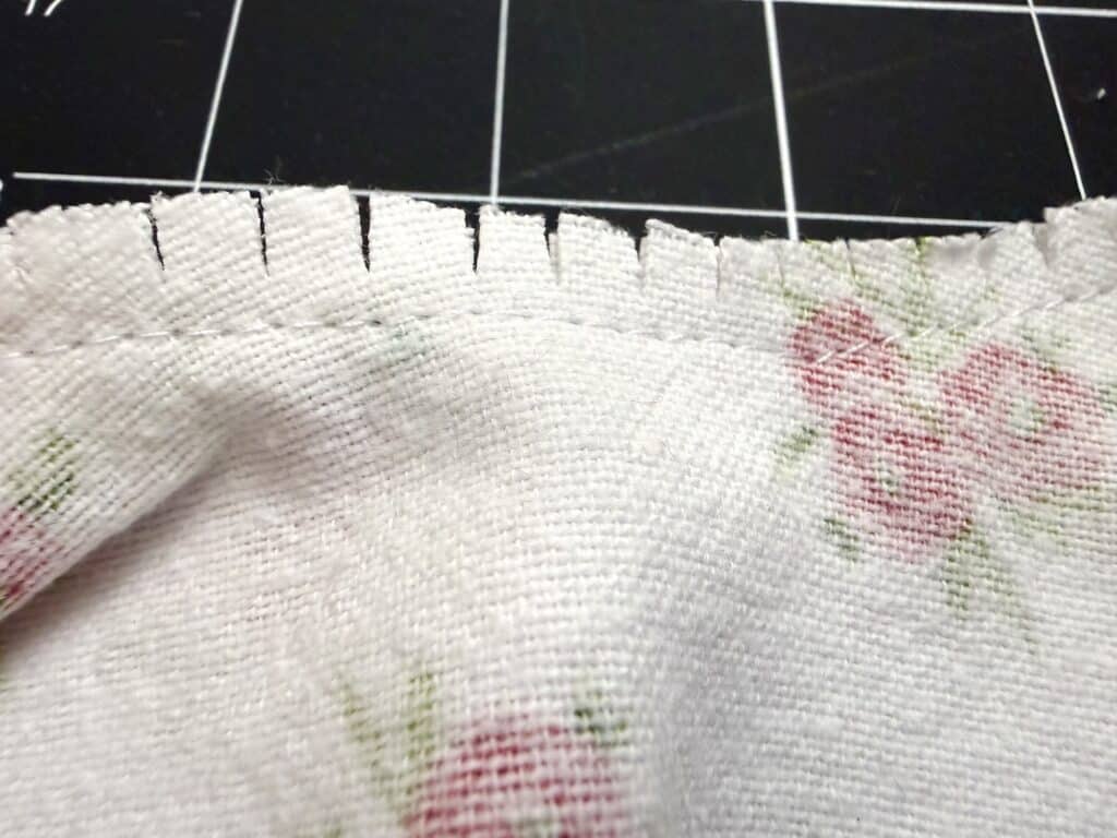 Close-up of a piece of white fabric with a floral pattern, showing notched cuts along the seam edge, placed on a black grid cutting mat.