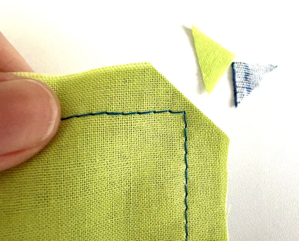 A hand holds green fabric with blue stitching along the edges; two small fabric triangles are cut off and placed nearby on a white surface.