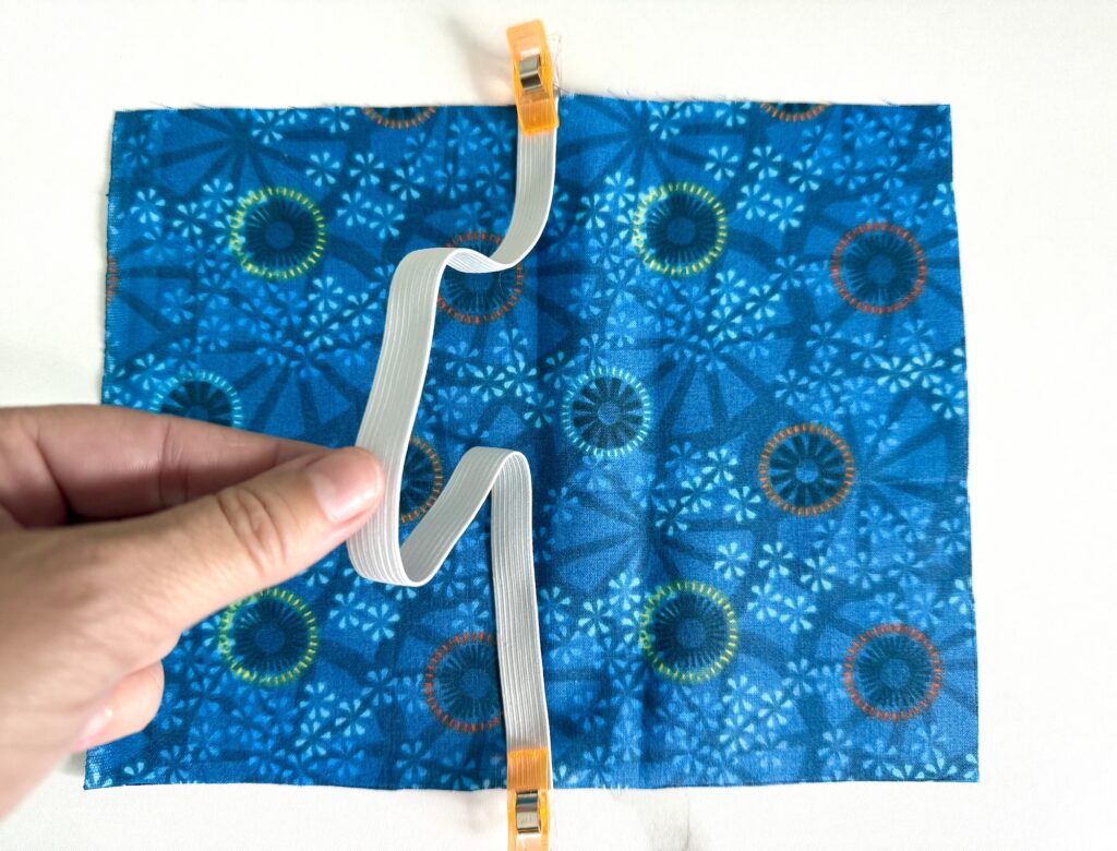 A hand holds a flat white cable clipped to a blue patterned fabric square with two orange clips, one on each side.