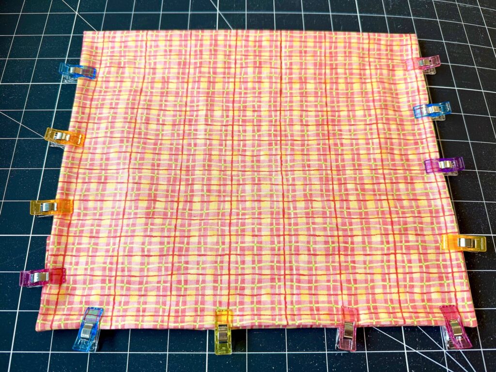 A rectangular piece of pink plaid fabric is held at the edges with colorful clips, placed on a black grid cutting mat.
