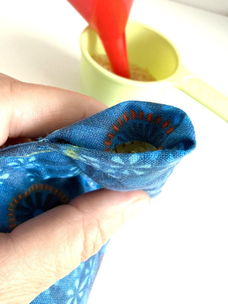 A hand holds a blue patterned fabric pouch with an opening while a yellow measuring cup and red funnel are visible in the background.