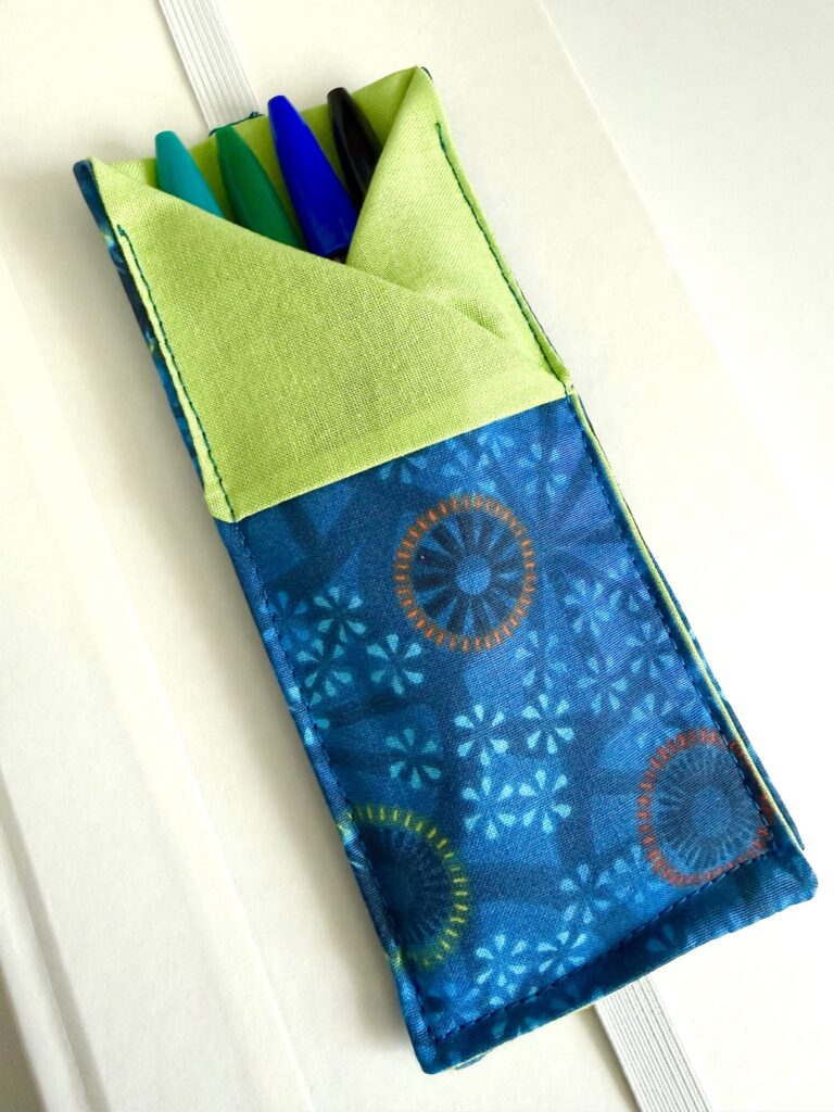 A blue and green fabric pen holder containing four pens is placed on a white surface.