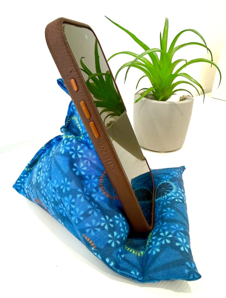 A smartphone in a brown case rests on a blue fabric stand with a plant in a white pot in the background.