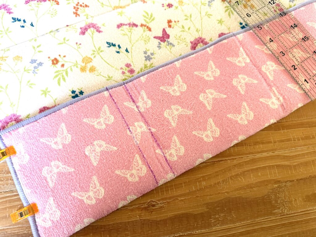 A pink fabric with white butterfly prints is measured with a clear ruler on a wooden surface. A floral fabric is partially visible above.