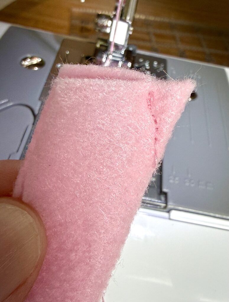 A close-up of a hand holding a piece of pink felt with a visible seam, in front of a sewing machine.