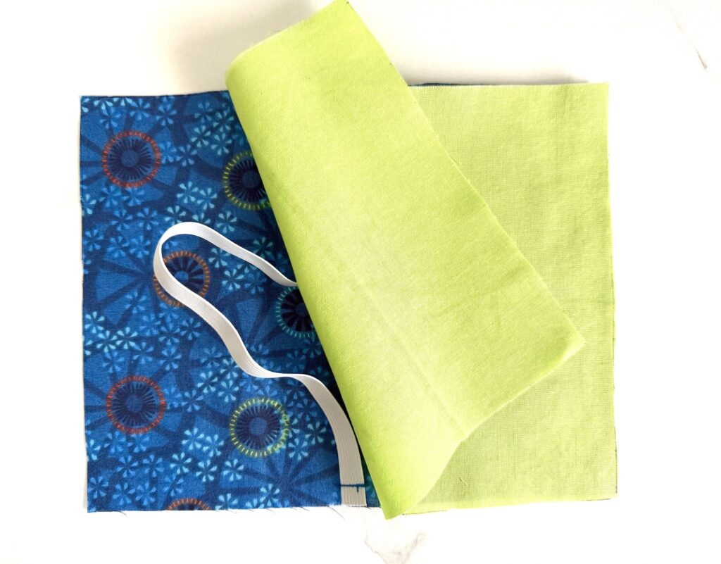 Two fabric squares, one blue with patterns and one solid light green, are stacked with a white elastic strip placed on top.