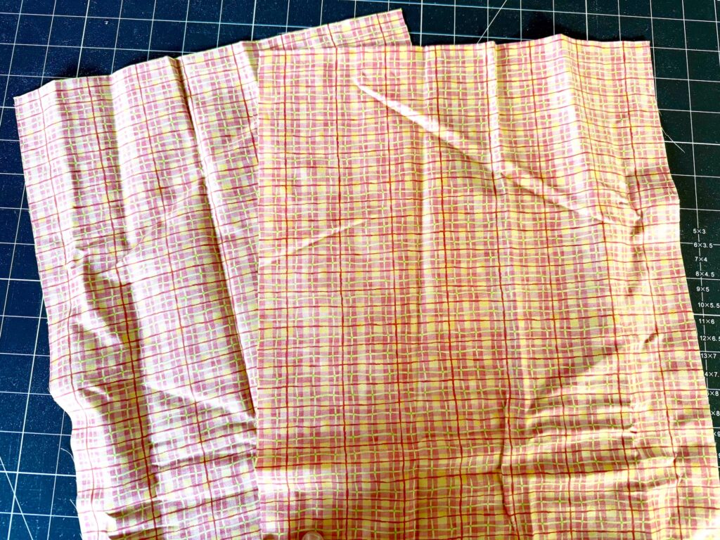 A piece of light pink fabric with a multicolored plaid pattern is laid out on a cutting mat with a visible grid.